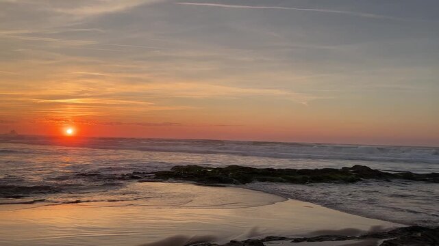 Intersecting waves and backwash flow create crisscrossing water patterns over wet sand at sunset on Atlantic beach. Rocks in water and distant coastal cliffs complete the calm seascape