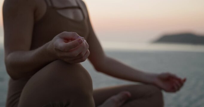 Crop view of young woman meditates in padmasana pose on dusk morning sea beach. Lady in sportswear holds hand in traditional mudra as rests on sunrise coast
