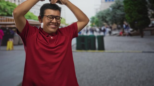 Man smiling and making a heart with hands while arms raised in a street market with souvenir stalls and trees visible; joy celebration.