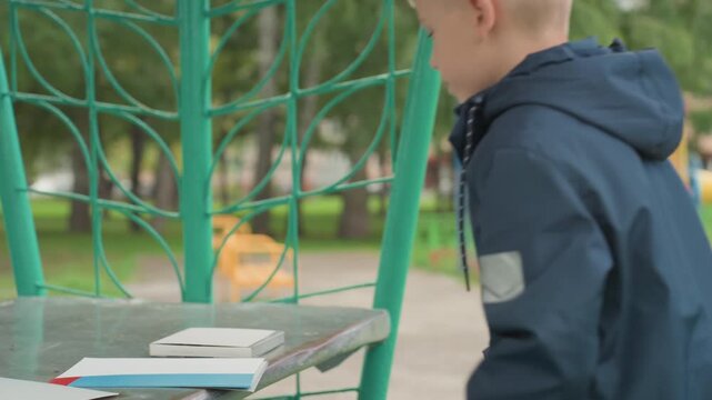 Boy places notebooks on bench near playground while woman observes, arranging papers and pencil, organizing backpack and supplies before school, focused independent routine and quiet preparation