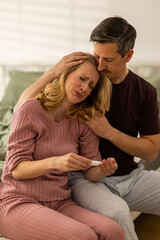 Obraz premium Couple Sits Together on Bed as Woman Looks at Pregnancy Test With Worried Expression in Modern Room
