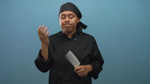 Man chef in black uniform holding a cleaver and touching his lip with a finger in a blue studio setting wearing a headscarf and doublebreasted coat; contemplation.