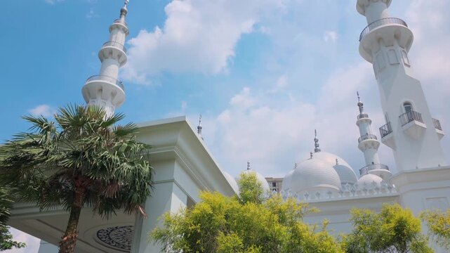 Depok, Indonesia (April 6, 2026): At-Thohir Mosque, A stunning view of a sacred white mosque with tall minarets set amidst lush trees and a bright sky, showcasing exquisite architecture and cultural
