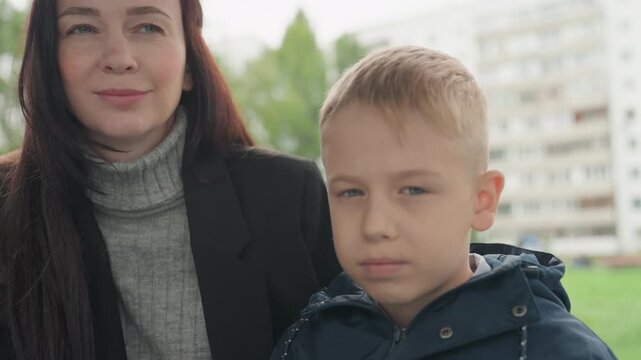 Mother and son prepare for school in residential park, woman in turtleneck adjusts boys jacket while apartment buildings blur in background, soft overcast light, calm morning atmosphere, emotional
