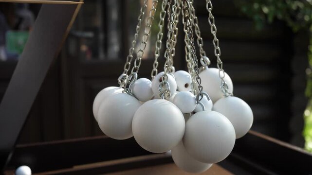 Female hand uses thin wooden stick to push cluster of matte white spheres suspended by silver metal chains within interactive outdoor leisure game.