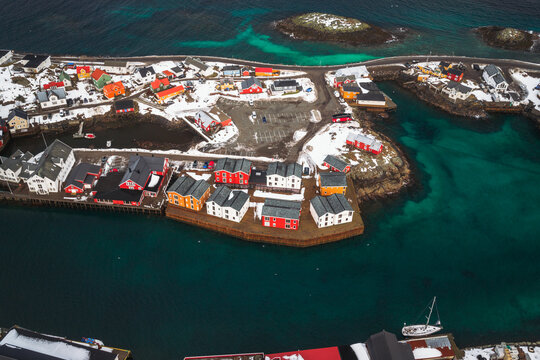 Aerial view of vibrant red and yellow buildings stand out against the snowy landscape and turquoise waters of Henningsvaer, Nordland, Norway.
