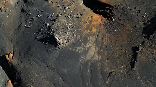 Top-down drone shot of dark volcanic ash and orange mineral streaks with scattered boulders, creating dramatic erosion patterns in Icelandic highland terrain.