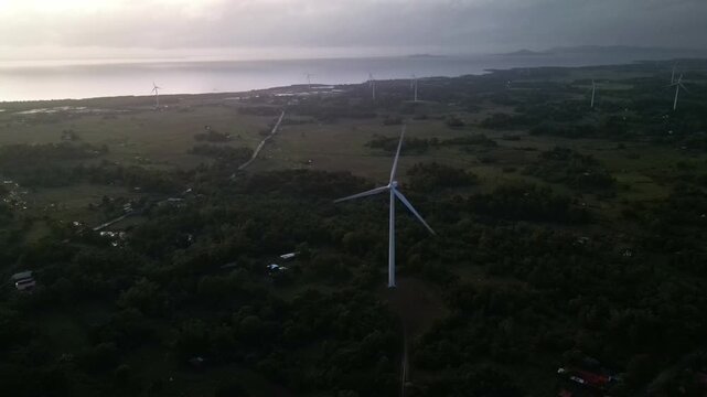 Aerial orbit of renewable energy wind turbines along lush, tropical island coastline at golden hour - Guimaras, Philippines