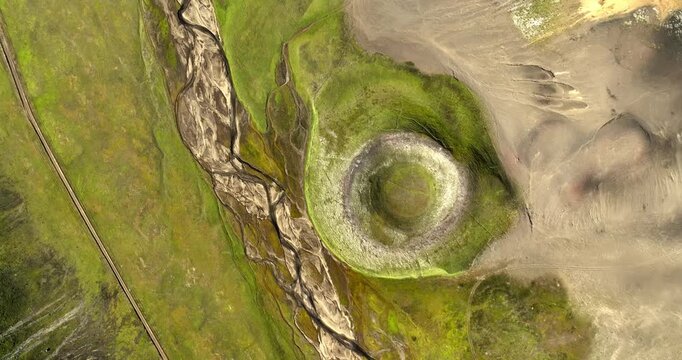 Top-down drone view of a green volcanic crater beside braided streams and ash plains, revealing striking natural patterns in a remote geothermal landscape.