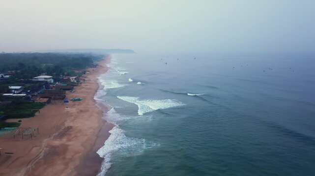 Coastal view of Calangute Beach at dawn, showing golden sand, breaking waves, and fishing boats on calm water. Residential buildings line the shore under soft morning light in Goa, India.