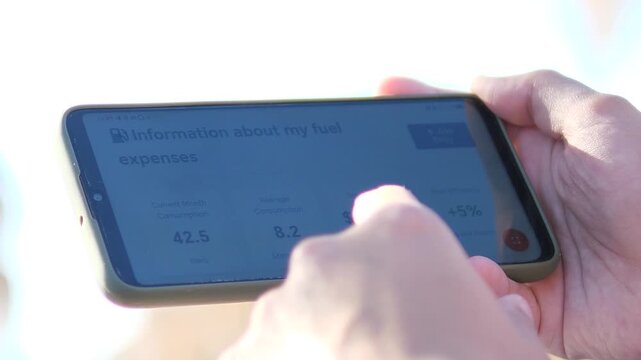 Car owner monitoring fuel efficiency on mobile device. POV shot of a modern app interface showing vehicle fuel analytics and monthly reports. Ideal for smart car reviews, tech promos, and software