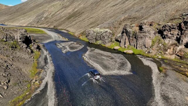 Drone aerial of a 4x4 SUV fording a braided river across gravel bars in Iceland&rsquo;s highlands canyon, with rocky cliffs and rugged mountain terrain.