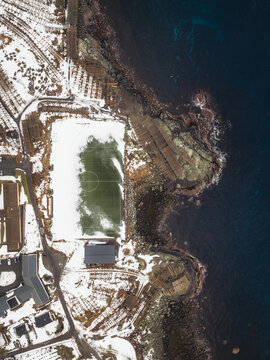 Aerial view of a snow-dusted landscape meeting the dark waters, where a soccer field stands as a bright green island, Reinebringen, Nordland, Norway.