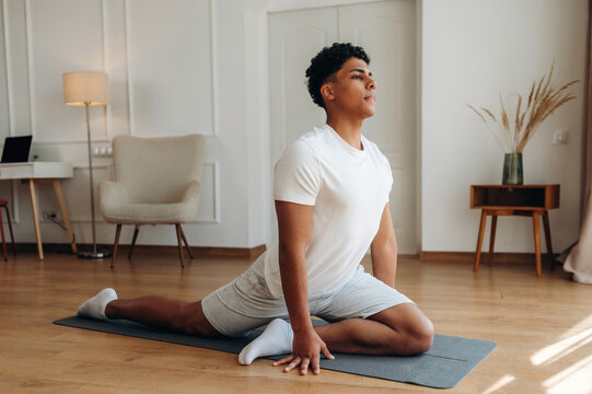 Young male practicing yoga in a modern indoor setting, performing a pigeon pose on a mat with wooden flooring and minimalistic decor in the background