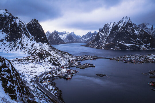 Aerial view of dramatic, snow-dusted peaks embracing the still, dark waters dotted with tiny, vibrant villages under a brooding sky, Reinebringen, Nordland, Norway.
