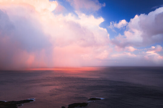 Aerial view of the vast, dark blue sea reflecting the fiery sunset beneath a blanket of pink-tinged clouds, Reinebringen, Nordland, Norway.