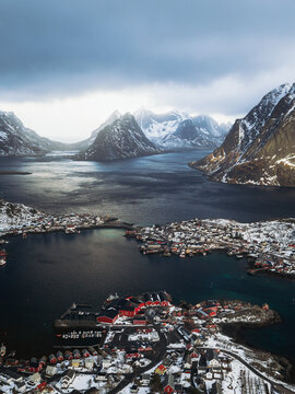 Aerial view of Reinebringen's village nestled amid frigid waters, jagged snow-capped peaks under brooding skies, Reinebringen, Nordland, Norway.