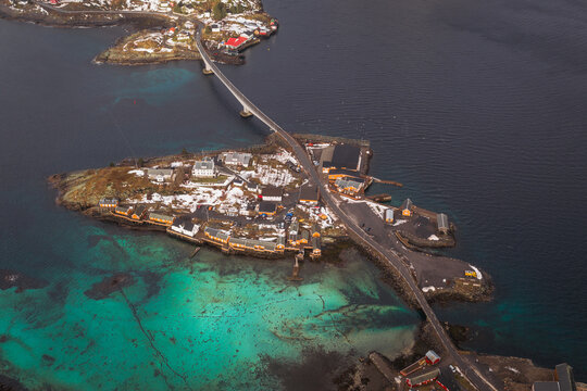 Aerial view of a bridge connecting islands, reflecting the turquoise waters below, with snow-dusted buildings adding contrast, Sakrisoya, Nordland, Norway.