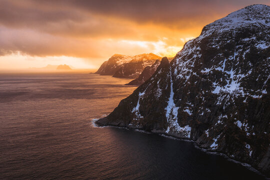 Aerial view of jagged, snow-dusted peaks plunging into the slate-blue sea under a fiery sunset sky, Nordland, Nordland, Norway.