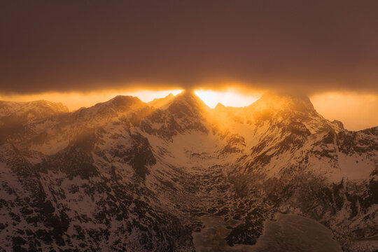 Aerial view of sun rays piercing through the clouds, illuminating the snow-capped peaks with a golden glow, creating a dramatic contrast against the dark, shadowed valleys, Nordland, Norway.