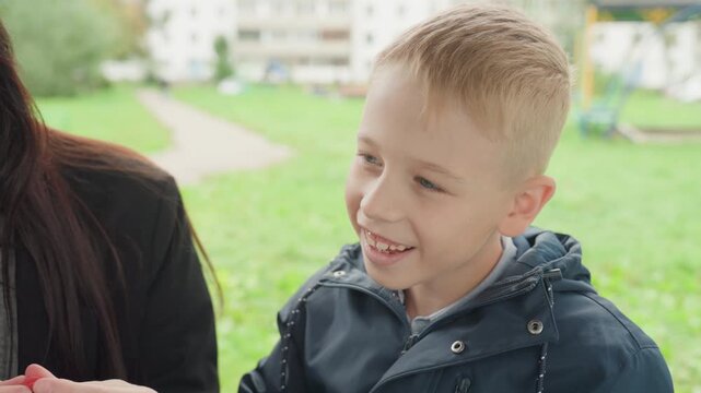 Mother and son preparing back to school, outdoor park bench interaction with jacket and stickers, joyful laughter, gentle caring touch, closeup expressions showing trust and bonding, morning routine