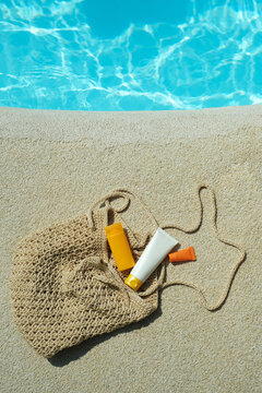 Sunscreen and lotion bottles rest on sandy ground next to a woven bag under direct sunlight near the pool. Summer skincare essentials for healthy and protected skin. top view