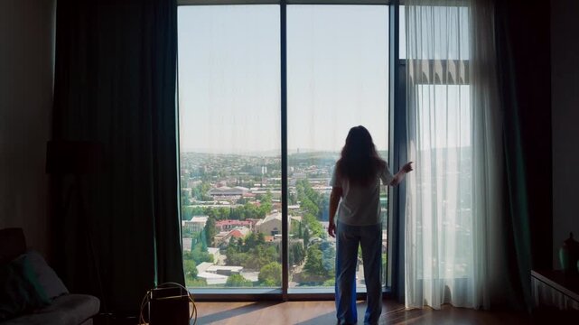 Rear view of a woman in pajamas walking to a panoramic window in a stylish apartment. She closes the sheer curtains, obscuring the bright city view and creating a cozy atmosphere