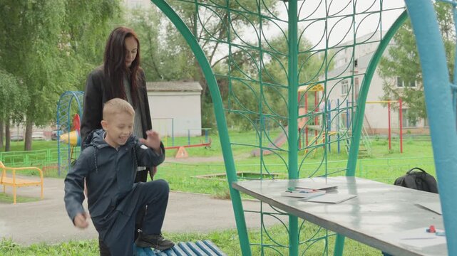 Playground stumble during back to school preparation caucasian mother comforts boy, helps him up after trip, checks for scrapes and guides back to bench for rest urban park setting with playground