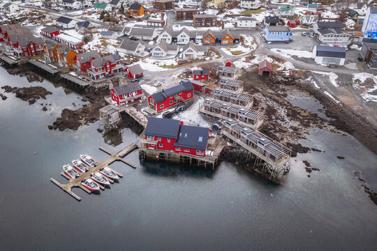 Aerial view of striking red buildings contrast against the muted greys of the surrounding landscape and the cold, still waters, Ballstad, Nordland, Norway.