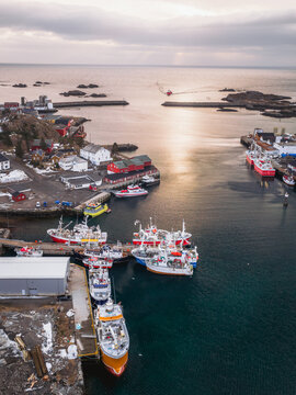 Aerial view of boats nestled in the harbor, vibrant against the deep blue water, with red buildings dotting the shoreline, Ballstad, Nordland, Norway.