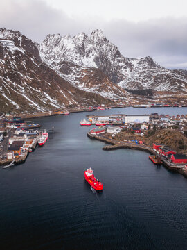 Aerial view of a deep blue fjord embraced by rugged, snow-dusted mountains and dotted with red buildings, the scene punctuated by a bright red ship, Ballstad, Nordland, Norway.