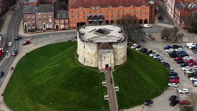 Aerial drone footage of Clifford Tower in the city of York in Yorkshire UK showing circular structure grass mound surrounding roads and car park captured directly from above in the spring time