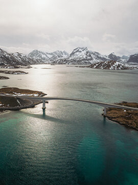 Aerial view of a sleek bridge elegantly spans the turquoise waters, contrasting with the rugged, snow-dusted mountains in the distance, Flakstadbruene, Norway.