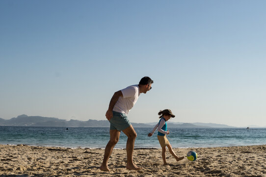 A wide shot of a young girl in a blue rash guard kicking a colorful ball while her father watches and plays along on a sunny sandy beach.