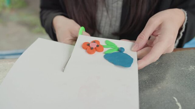 Asian woman trimming paper with scissors prepare back to school art poster featuring foam floral sticker closeup hands aligning edges on desk focused tutorial classroom project vibrant supplies final