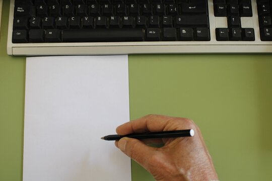 Primer plano de una mano sosteniendo un l&aacute;piz sobre una hoja en blanco, lista para escribir o dibujar, sobre un escritorio con teclado al fondo en la oficina del trabajo. Ideal para texto y dise&ntilde;o.   