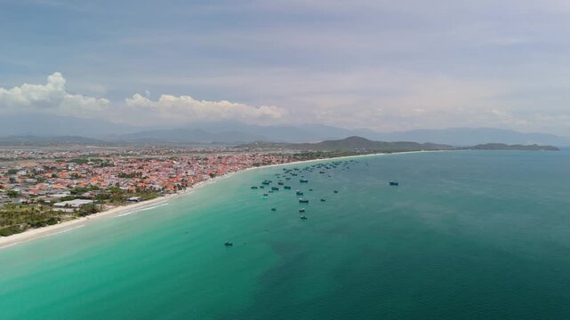 Watch as fishing boats float on the water near Dok Let Beach in Vietnam. White sand stretches along the coast, and colorful houses line the shore. Sunbathers relax under umbrellas