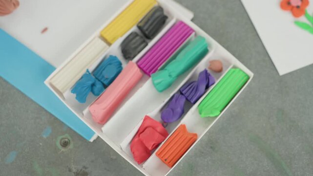 Colorful modeling clay set back to school prepare with open organizer on classroom desk, sorted vibrant sticks in multiple colors, small paper flower beside kit, textured work surface with clay
