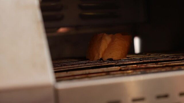 A slice of bread moves slowly along a conveyor toaster belt inside a breakfast serving station where prepared items are heated before guest selection. Food serving.