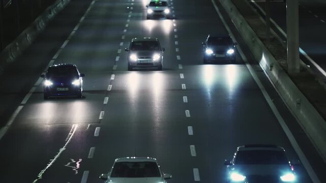 MADRID, SPAIN - JANUARY 13, 2026: Multiple car driving toward camera on a multilane urban highway at night, bright headlight illuminating wet asphalt, concrete barrier on both side
