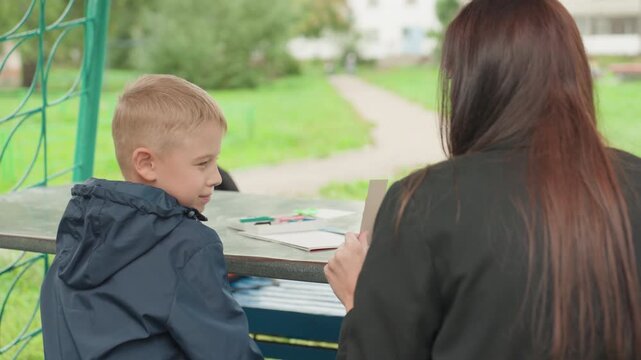 White mother comforts son at gazebo preparing for back to school, sharing handmade card and gentle hug, outdoor craft table, calm park path in background, affectionate reunion and support