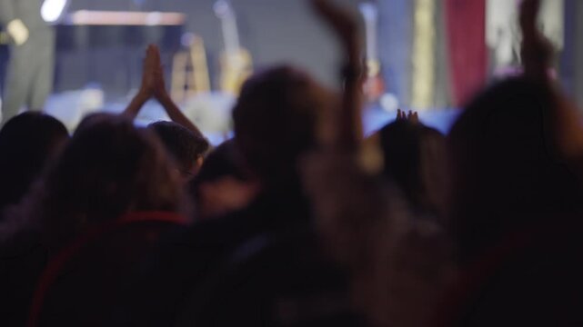 Shot from behind, an audience in silhouette watches a show. The crowd energy builds as they begin to clap, cheer, and raise their hands in applause for the performance on a brightly lit stage