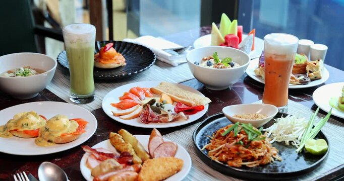 Luxurious breakfast buffet spread on a table by a window. Abundant international cuisine, fresh fruits, and beverages. Upscale hotel dining, slow motion.
