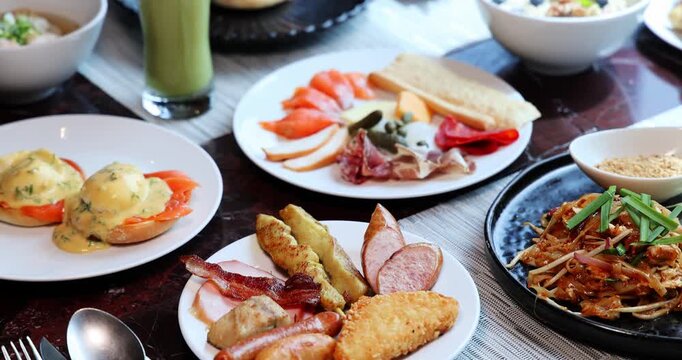 Abundant breakfast buffet featuring Eggs Benedict, sausages, bacon, Pad Thai, and smoked salmon on a dark marble table. Luxurious dining experience. High-angle, static shot.