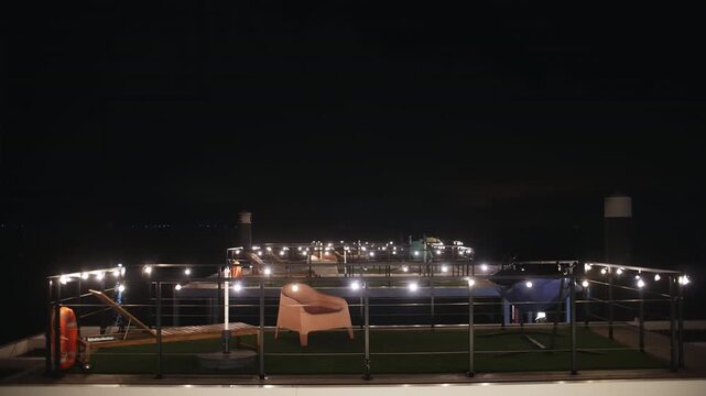 A tranquil night scene on an illuminated outdoor rooftop deck, featuring cozy lounge furniture, string lights, artificial grass, and a distant waterfront view