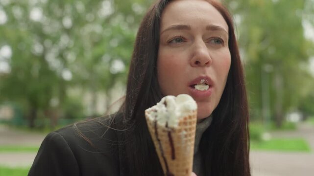 Caucasian woman holding ice cream cone outdoors, slow contemplative bite of waffle cone with vanilla cream, light melting on fingers, park trees and soft green background, wearing dark coat