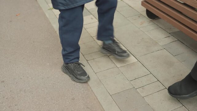 Legs and shoes of student standing near bench on paved sidewalk, casual sneakers with scuffed toes, coat hem visible, waiting, prepare for back to school, candid outdoor moment capturing footwear
