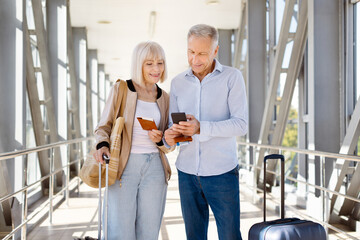 Mature couple standing in sunlit airport corridor, man using smartphone while woman holds passport and boarding pass, both smiling at screen, ready for travel © Anastasiya