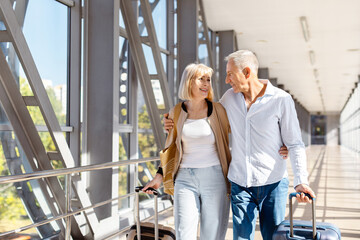 Mature couple walking together through modern airport corridor with rolling suitcases, happy aged...