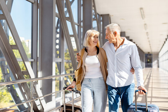 Mature couple walking together through modern airport corridor with rolling suitcases, happy aged spouses smiling at each other in casual travel attire